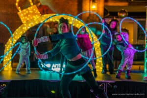 A performer uses glowing LED hula hoops in an engaging act on stage, surrounded by children with hoops, under colorful lights in Salem, Massachusetts.