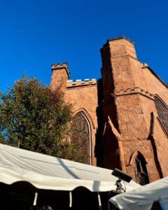 Close-up of the Salem Witch Museum in Salem, Massachusetts, showcasing its Gothic-style architecture with pointed towers and arched windows, framed by a bright blue sky. A white tent and nearby foliage are visible in the foreground.