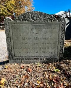 The gravestone of Mary Masury, wife of William Masury, in the Old Burying Point Cemetery in Salem, Massachusetts. The stone is engraved with a cherubic face with wings at the top, surrounded by ornate floral carvings. The inscription marks her passing on May 17, 1744, at the age of 27. The gravestone is set in a historic cemetery under a bright blue sky, surrounded by fallen autumn leaves.