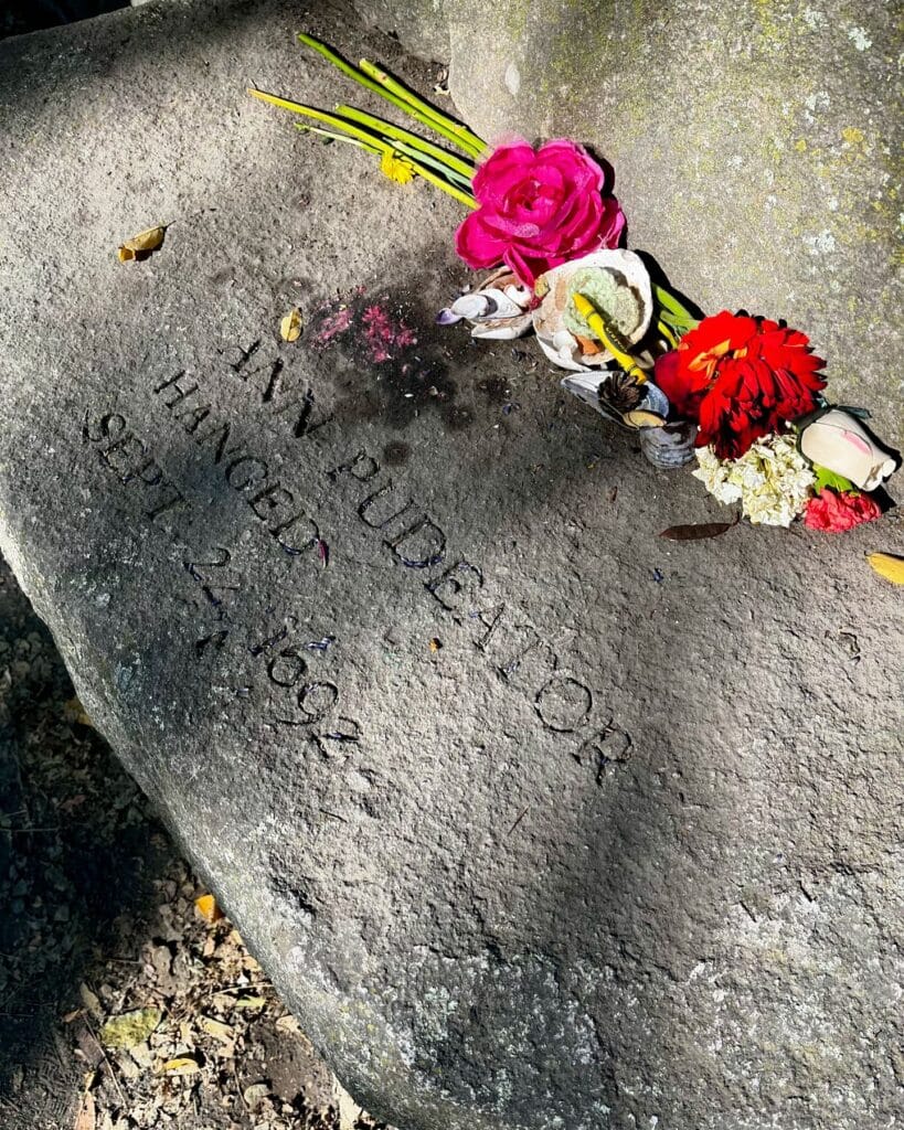 A memorial stone at the Salem Witch Trials Memorial in Salem, Massachusetts, honoring Ann Pudeator, engraved with her name and the date of her execution, September 22, 1692. Bright flowers and seashells rest on the stone.