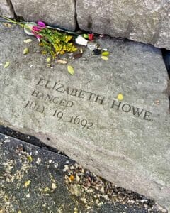 A memorial stone in Salem, Massachusetts honoring Elizabeth Howe, a victim of the Salem Witch Trials, engraved with her name and the date of her execution, July 19, 1692. Flowers are placed on the stone, surrounded by fallen leaves.