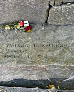 A memorial stone at the Salem Witch Trials Memorial in Salem, Massachusetts, honoring George Burroughs, engraved with his name and the date of his execution, August 19, 1692. Flowers and small tokens are placed on the stone.