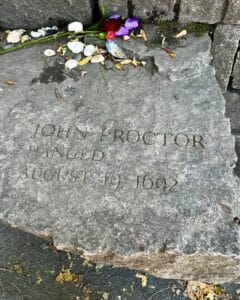 A memorial stone at the Salem Witch Trials Memorial in Salem, Massachusetts, honoring John Proctor, engraved with his name and the date of his execution, August 19, 1692. Flowers, shells, and small tokens rest on the stone.