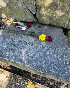 A memorial stone at the Salem Witch Trials Memorial in Salem, Massachusetts, commemorating Margaret Scott, engraved with her name and the date of her execution, September 22, 1692. Flowers, including a yellow bloom, are placed on the stone.