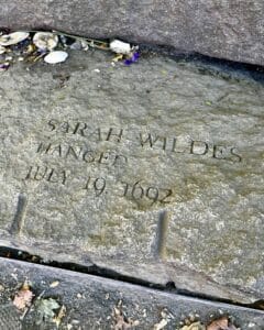 Engraved memorial stone for Sarah Wildes at the Salem Witch Trials Memorial in Salem Massachusetts, inscribed with her name, the date July 19, 1692, and surrounded by offerings like flowers and seashells.
