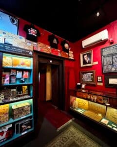 Inside the Salem Witch Board Museum, a wall features shelves with Ouija board-themed merchandise, including t-shirts and books. Nearby, a display case showcases vintage Ouija boards, and framed historical information adorns a vivid red wall.