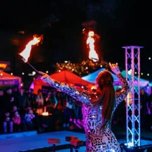 A fire dancer in a reflective costume performs with flaming batons, surrounded by colorful tents and a captivated crowd during a nighttime event in Salem, Massachusetts.