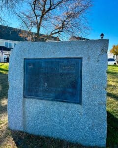 A granite monument in Salem, Massachusetts, featuring a plaque commemorating the Salem Frigate, also known as the Essex, with a tree and historic buildings in the background.