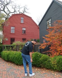 The red colonial-style building is Nathaniel Hawthorne's Birthplace in Salem, Massachusetts, with a person standing on a brick pathway in front of green hedges.