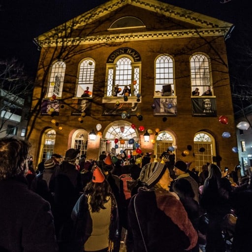 A crowd gathered outside the brightly lit Old Town Hall in Salem Massachusetts, watching people lean out of open windows and enjoying a festive evening atmosphere.