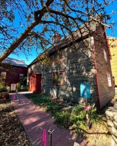 The Old Burying Point Charter Street Cemetery Welcome Center in Salem Massachusetts, featuring a colonial-style structure with red trim, a brick pathway, and autumnal trees.
