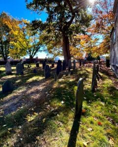 Gravestones in a historic Salem Massachusetts cemetery surrounded by vibrant autumn leaves, sunlight streaming through tall trees, and a peaceful grassy setting.