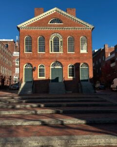The Old Town Hall in Salem, Massachusetts, a historic red brick building with arched windows and green double doors. Stone steps lead up to the entrance, with long shadows cast across the brick plaza.