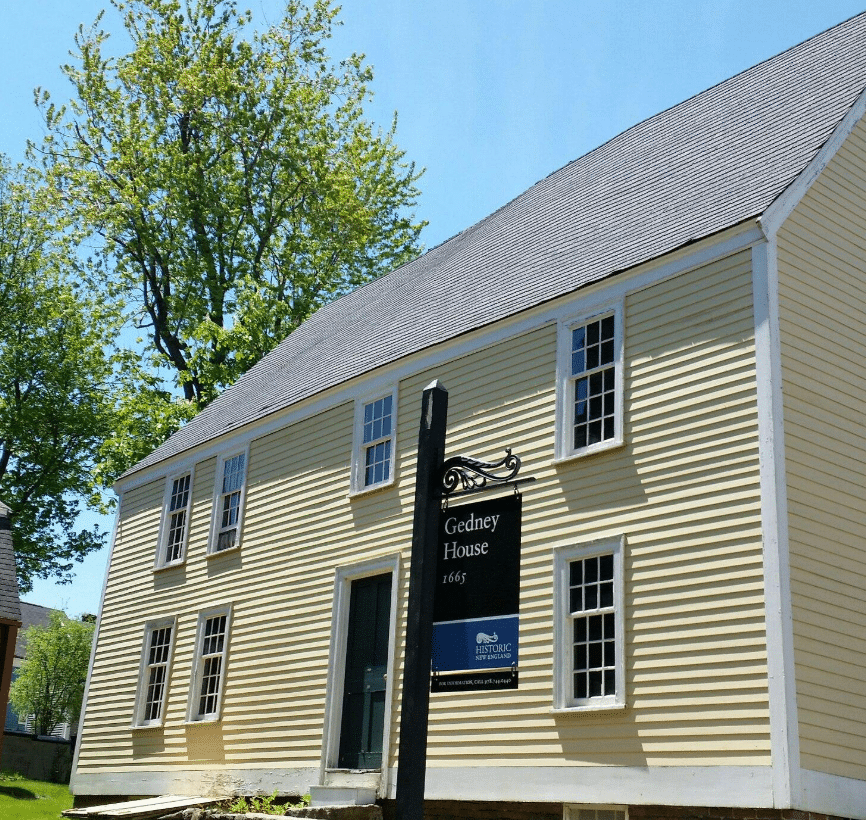 This image features the Gedney House, a historic First Period structure built in 1665, located in Salem, Massachusetts. The yellow clapboard exterior, steeply pitched roof, and white trim reflect its colonial architectural style. Prominently displayed in front is a sign identifying the house as part of Historic New England, the organization that preserves and manages the site. Surrounded by trees and bathed in sunlight, the Gedney House stands as a well-preserved example of 17th-century craftsmanship and history, offering visitors a glimpse into early colonial life.