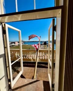 A view from the Salem Custom House in Salem, Massachusetts, showing open balcony doors, the American flag blowing in the wind, and Salem Harbor under a clear blue sky.