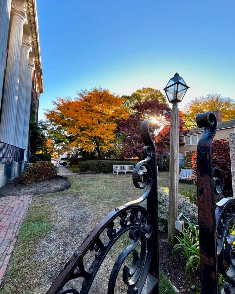 Ornate iron gate framing the view of the Andrew-Safford House garden in Salem Massachusetts, with benches, colorful autumn trees, and sunlight glowing through a vintage lamp.