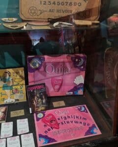 A display at the Salem Witch Board Museum featuring Ouija boards, including a pink "Mystifying Oracle" set and vintage wooden boards. A book titled "Creepover" and various planchettes are also showcased.