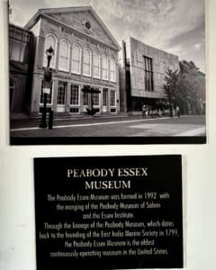 Black-and-white image of the Peabody Essex Museum in Salem, Massachusetts, featuring East India Marine Hall alongside a historical sign describing the museum's founding and lineage.