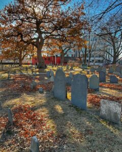 Old gravestones and a large tree with autumn leaves at Old Burying Point Cemetery in Salem, Massachusetts. Sunlight filters through the branches, casting long shadows on the ground covered in fallen leaves.