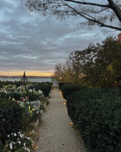 A garden path at the House of the Seven Gables in Salem, Massachusetts, leads to a waterfront view at sunset. Flowers and hedges line the walkway, with soft lights guiding the way.