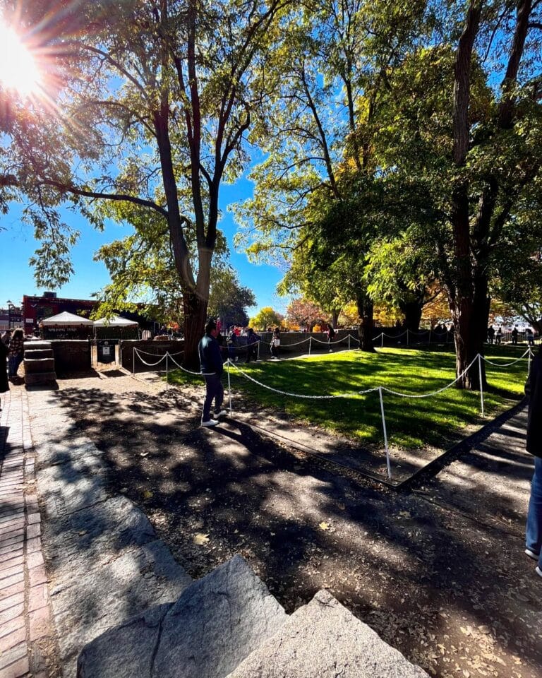 A sunny view of the Salem Witch Trials Memorial in Salem, Massachusetts, showing green grass, tall trees, and pathways lined with white ropes. Visitors walk through the serene space under the bright blue sky.