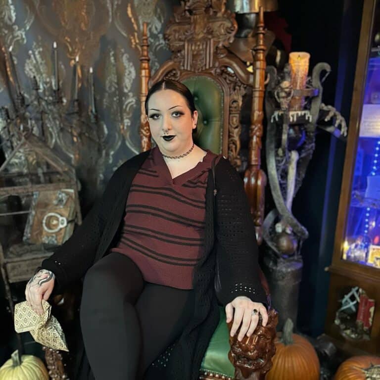 A person dressed in dark, gothic clothing sits confidently on an ornate wooden throne, surrounded by Halloween decorations. The chair is green and intricately carved, and the setting features spooky elements like pumpkins, cobwebbed candleholders, and vintage curiosities, creating a mysterious atmosphere.