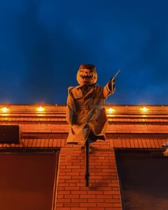 A spooky pumpkin head figure wearing a trench coat and pointing a stick stands above a brick building in Salem, Massachusetts, illuminated by orange lights at night. The dark sky adds to the eerie atmosphere, creating a classic Halloween vibe.