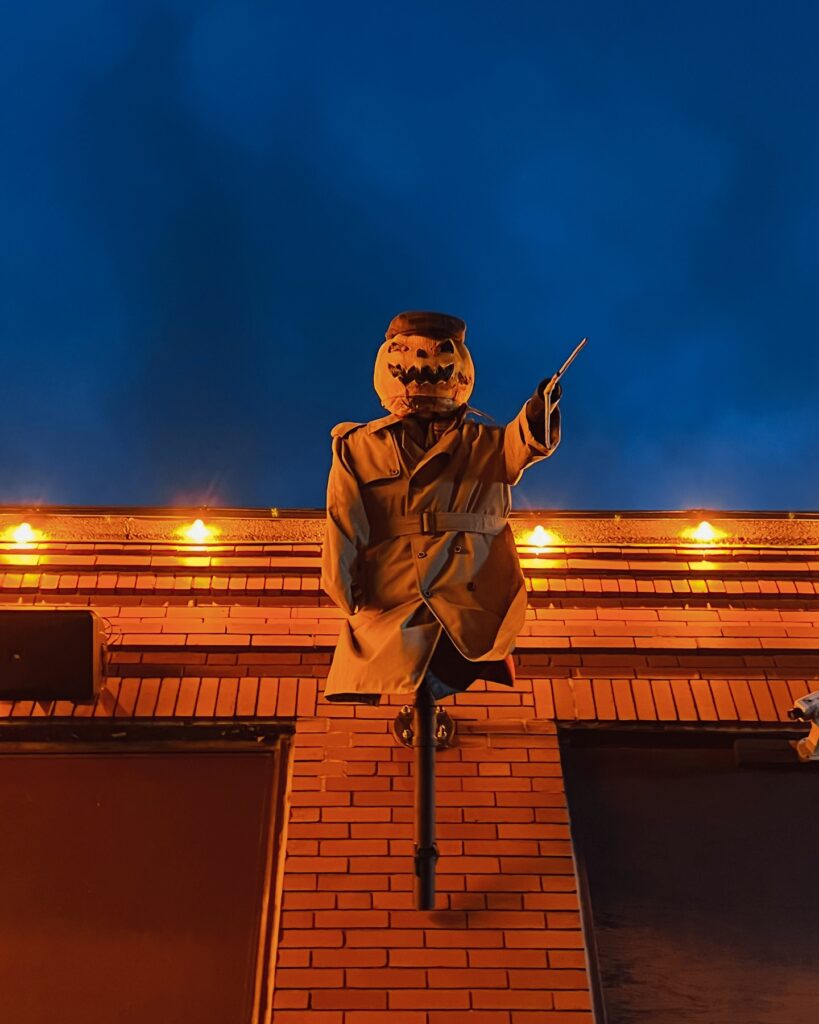 A spooky pumpkin head figure wearing a trench coat and pointing a stick stands above a brick building in Salem, Massachusetts, illuminated by orange lights at night. The dark sky adds to the eerie atmosphere, creating a classic Halloween vibe.