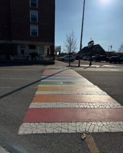 A rainbow-colored crosswalk with cracked paint stretches across the street in Salem, Massachusetts. A brick building and parked cars are visible in the background under a clear sky.