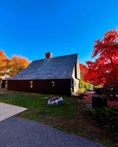 A rear-angle view of the historic John Ward House in Salem, Massachusetts, featuring its dark wood siding and shingled roof, framed by vibrant red and orange autumn trees against a clear blue sky.