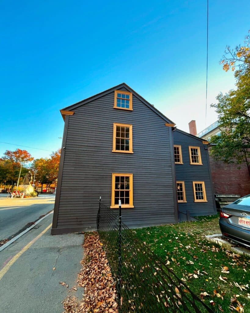 A rear side view of the Daniel Bray House in Salem, Massachusetts, featuring dark wood siding with golden trim, a black fence along the sidewalk, and scattered autumn leaves on the grass.