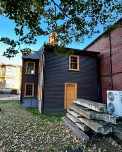 The rear view of the Daniel Bray House in Salem, Massachusetts, showing its dark wood siding, contrasting golden trim, and stacked stone beams nearby.