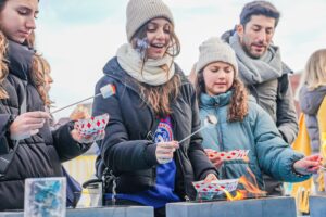 A group of attendees enjoying the activity of roasting marshmallows over open flames during the Frozen Fire Festival in Salem, Massachusetts, while bundled up in warm winter clothing.