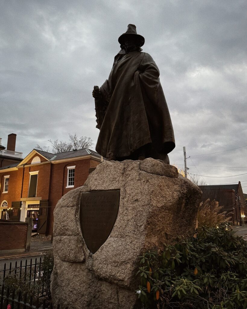 The Roger Conant statue in Salem, Massachusetts, features a bronze figure in a long cloak and hat, standing atop a large stone base. A plaque is affixed to the front of the stone, with historic buildings in the background.