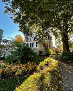 Historic Ropes Mansion in Salem, Massachusetts, partially shaded by a large tree with autumn leaves and garden plants along a gravel path.