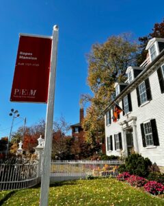 The Ropes Mansion in Salem, Massachusetts, with a red sign from the Peabody Essex Museum stating "Ropes Mansion, Built 1727–1729," set near a white picket fence, colorful gardens, and fall trees under a bright blue sky.