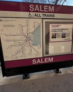 A sign at Salem Train Station in Salem, Massachusetts, displaying a commuter rail map and travel information. The sign reads "SALEM" and "ALL TRAINS," with the Massachusetts Bay Transportation Authority (MBTA) map below.