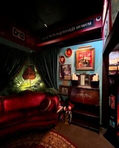 A warm and inviting corner of the Salem Witch Board Museum featuring a plush red velvet sofa, elegant green curtains, and vintage decor elements.