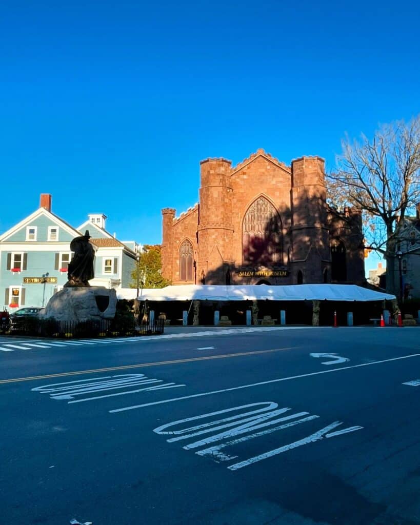 The Salem Witch Museum in Salem, Massachusetts, featuring its Gothic architecture, flanked by neighboring historic buildings and a statue in the foreground, under a bright blue sky.