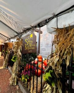 Sign of the Salem Witch Museum featuring a yellow witch graphic and the year 1692, surrounded by fall decorations, including pumpkins, cornstalks, and hay bales, partially covered by a white tent.