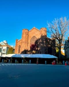 The Salem Witch Museum in Salem, Massachusetts, stands majestically under a vivid blue sky, adorned with seasonal decorations including cornstalks and hay bales. A white canopy stretches across the front of the building, with orange traffic cones marking the area.