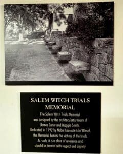 A display at the Salem Witch Trials Memorial featuring a historical black-and-white photo of the memorial's stone benches and a plaque with details about the memorial's dedication.