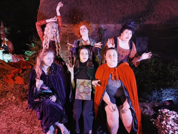 ix people dressed as the Sanderson Sisters from "Hocus Pocus" in Salem, posing dramatically with a rustic cabin in the background, illuminated by eerie lighting.