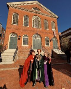 Three people dressed as the Sanderson Sisters from Hocus Pocus stand in front of the Old Town Hall in Salem, Massachusetts, a historic red-brick building with large arched windows and classic green double doors.