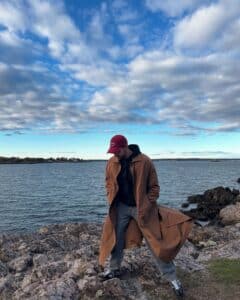 A man in a long brown coat and red cap stands on rocky terrain by Salem Harbor in Salem, Massachusetts. The ocean stretches behind him under a sky filled with clouds.