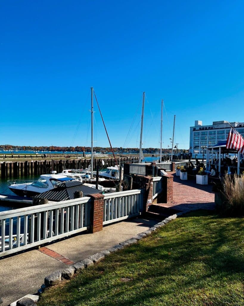 Boats docked at the marina in Salem Harbor, Salem, Massachusetts, surrounded by calm waters, a well-maintained walkway, and distant historic and modern buildings under a clear blue sky.