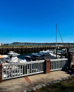 Boats docked at the marina in Salem Harbor, Salem, Massachusetts, with a clear blue sky and the distant shoreline framed by calm waters and a wooden railing.