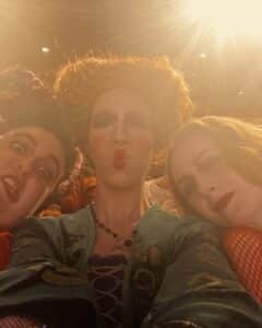 Three friends dressed as the Sanderson Sisters from Hocus Pocus taking a selfie under bright stage lights. Their costumes are detailed, with heavy makeup, red lips, and dramatic expressions, while the lighting creates a golden glow."