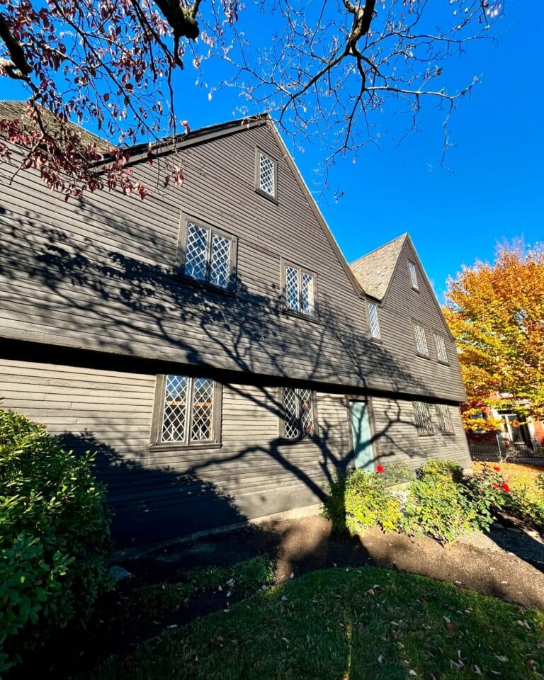 The historic John Ward House in Salem Massachusetts with its dark wood exterior, diamond-paned windows, and shadows of bare tree branches against a vibrant autumn background.