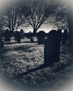 A black-and-white image of gravestones at Old Burying Point Cemetery in Salem, Massachusetts. Long shadows stretch across the grass, with bare trees framing the scene in the background.